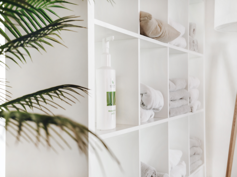 Regal mit weissen Handtüchern und Massage-Lotion in einem hellen, entspannten Behandlungsraum