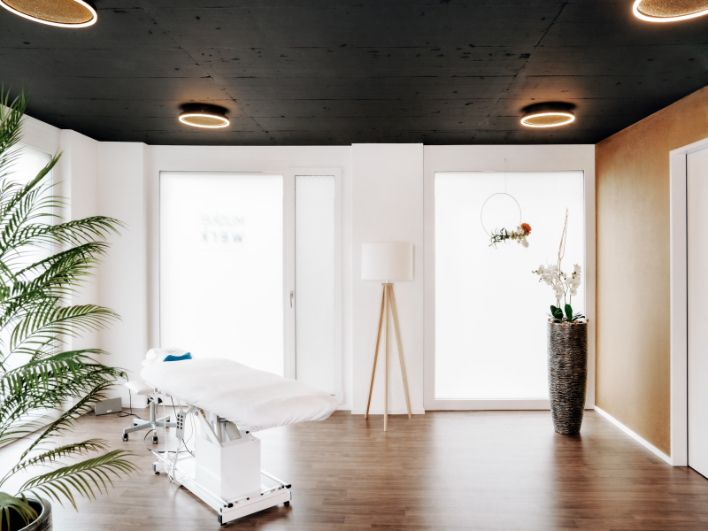 Moderner, heller Behandlungsraum mit Massageliege, dekorativen Pflanzen und stilvoller Beleuchtung